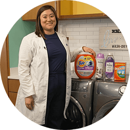 An Asian female wearing a white lab coat stands next to a washer and dryer. Several fabric care products are lined on top of the machines.