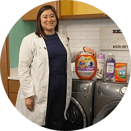 An Asian female wearing a white lab coat stands next to a washer and dryer. Several fabric care products are lined on top of the machines.