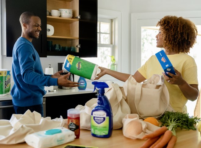 Woman and man are unpacking groceries in kitchen. Woman hands man a roll of Bounty paper towels.