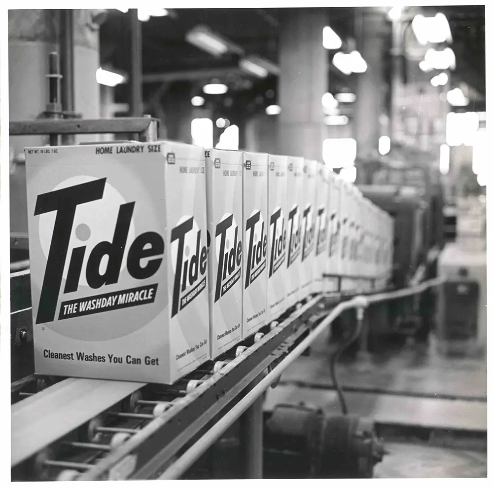 A black and white image of boxes of Tide move down the manufacturing line in 1965.