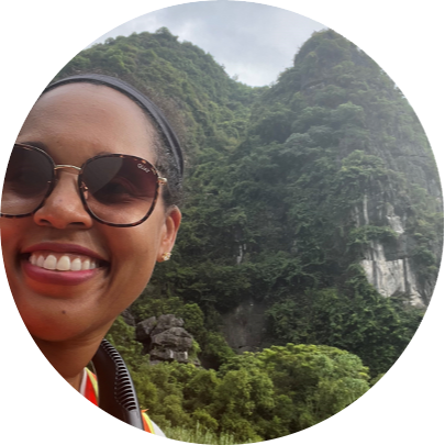 A black woman in sunglasses smiles as she stands in front of a large, tree-covered mountain in Vietnam.