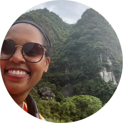 A black woman in sunglasses smiles as she stands in front of a large, tree-covered mountain in Vietnam.