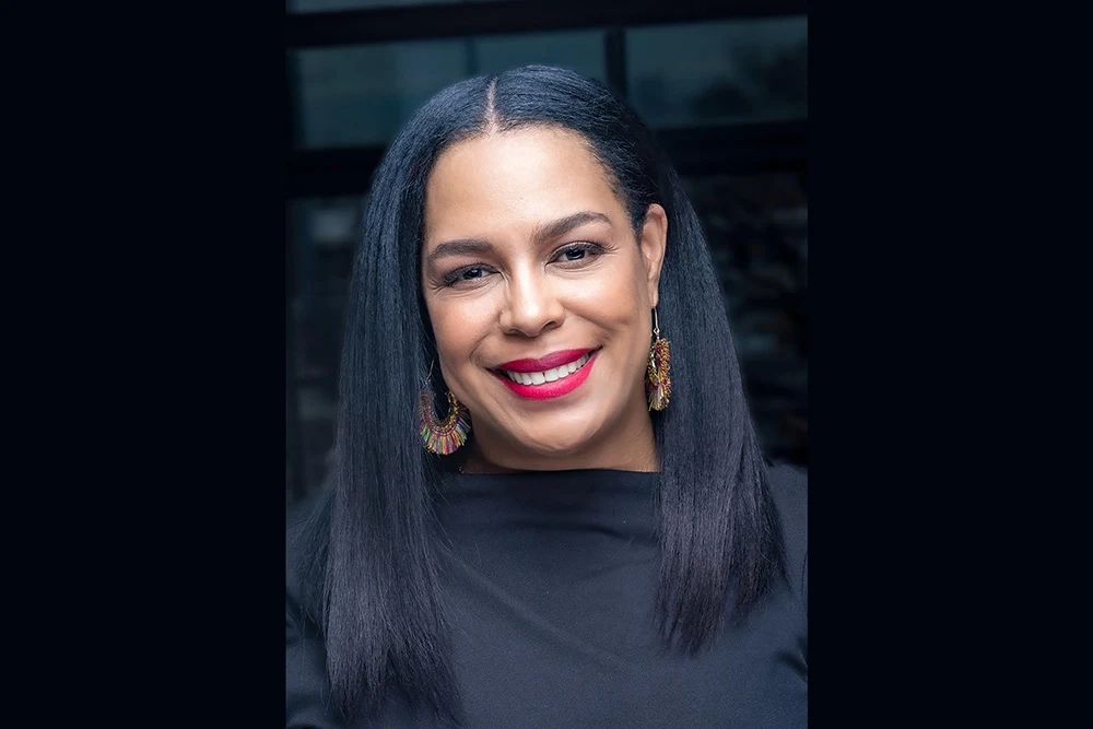 A black woman with long, dark straight hair wears a black blouse and large gold earrings as she smiles at the camera.
