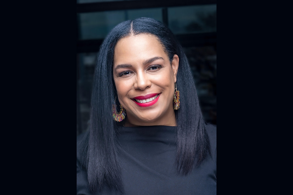 A black woman with long, dark straight hair wears a black blouse and large gold earrings as she smiles at the camera.