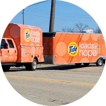 An orange truck and connected trailer has the Tide logo on them and says ‘loads of hope’.  