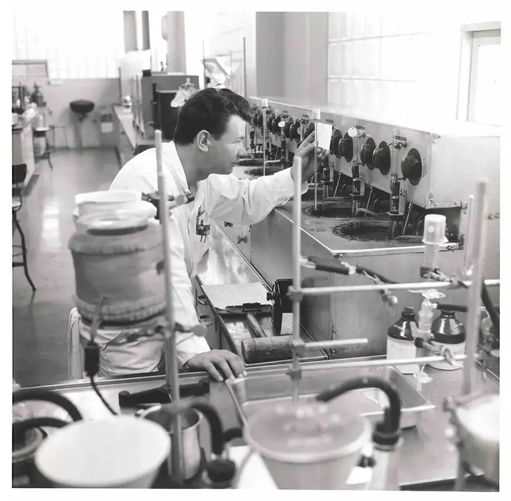 Black and white image of a man in a Tide lab taking measurements at a piece of equipment.