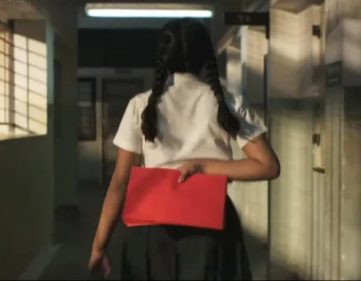 Girl in a school hallway from behind  with dark hair. She is carrying a bunch of red papers behind her back in her hand.