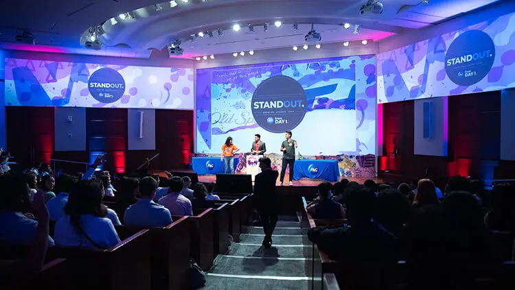 One woman and two men stand on an auditorium stage, engaging with the audience during a Q&A session.