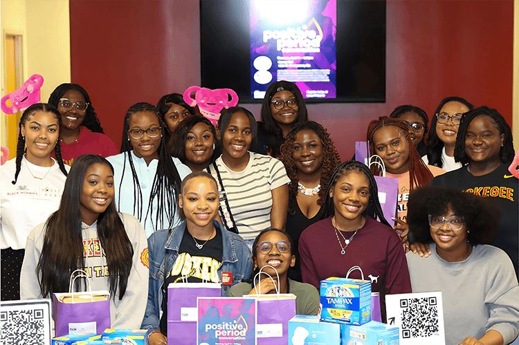 A group of seventeen young black college women gather and smile in front a table displaying Tampax period products and packaging.
