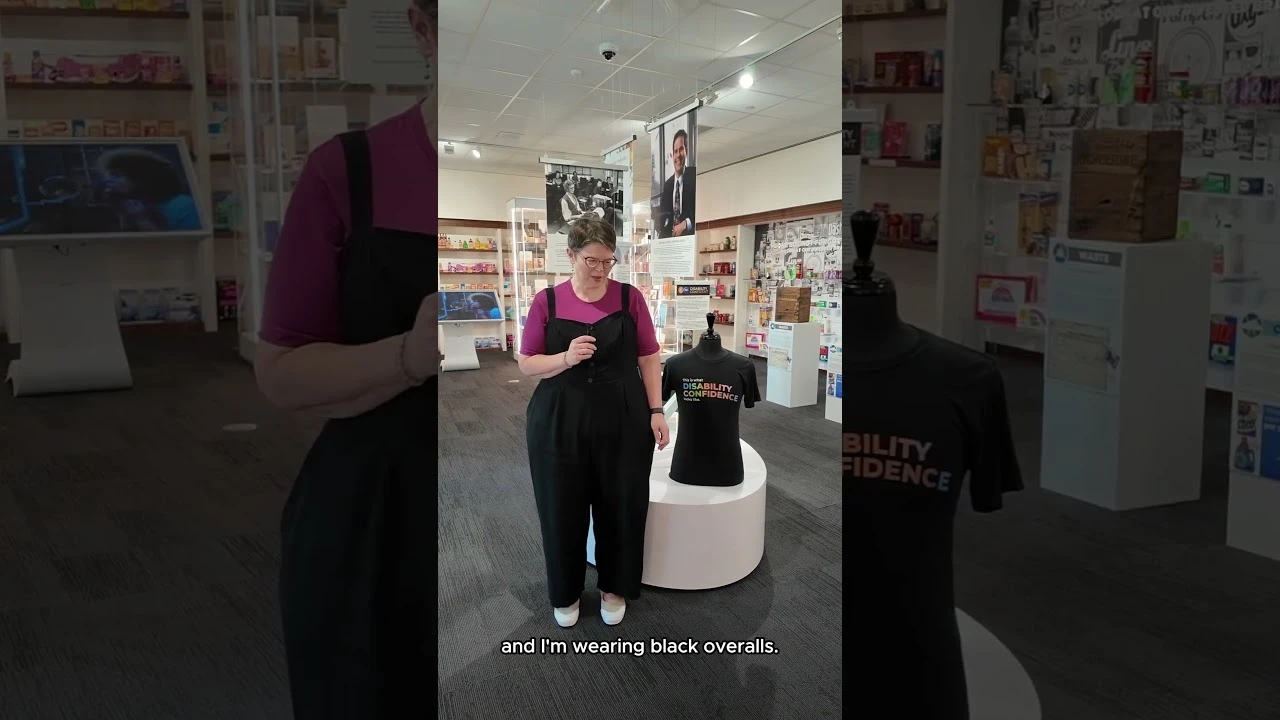 A woman with short brown hair and glasses talking into a small microphone about her experience as a caregiver. She wears a pink t-shirt and black overalls.
