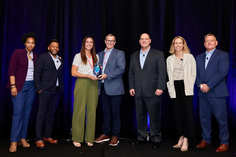 Four men and three women stand side by side on a stage with a dark backdrop. A man and woman in the center hold up a glass trophy.