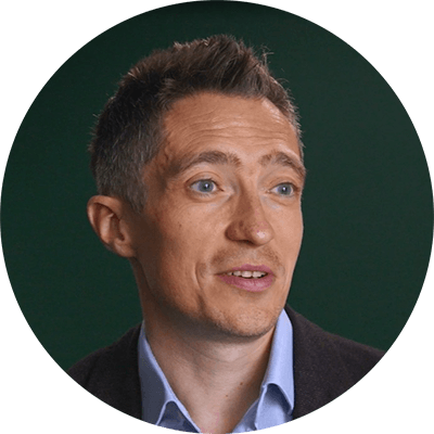 A man with short brown hear. He wears a light blue shirt and a black jacket.