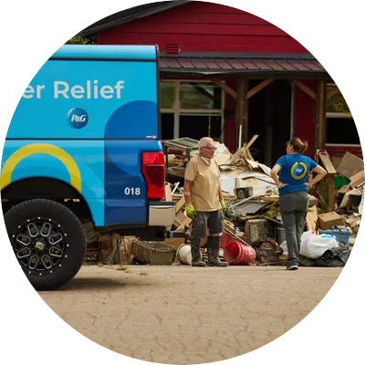 A blue truck branded with Walmart.org and P&G logos is parked in front of a flood-damaged building. Two people stand talking near piles of debris, broken wood, household items, and trash bags.
