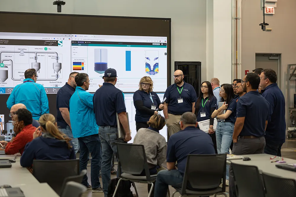 A group of about 15 SPARK participants in a classroom setting with an LED board in the background displaying manufacturing process components