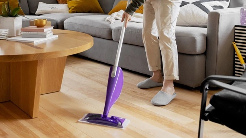 A person is cleaning the hardwood floors in a living room. They are only visible from the hip down, and they are using a purple Swiffer PowerMop.