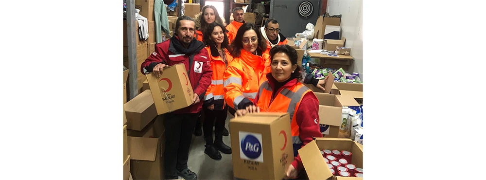 A group of people in orange safety vests inside a room filled with boxes, holding P&G and IFRC branded boxes of aid.