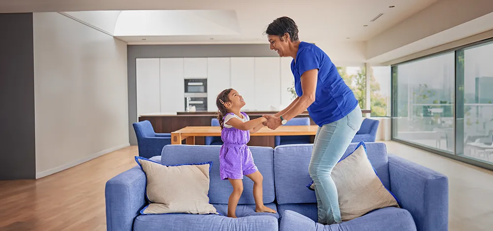 An older woman and a young girl smile and hold hands while they jump on a blue sofa in a bright, modern living room.