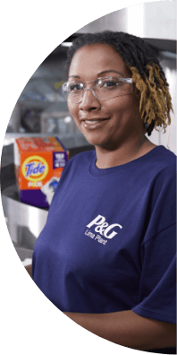  A woman wearing safety glasses and a blue t-shirt that says "P&G Lima Plant" smiles with a box of Tide pods in the background.