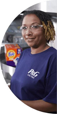 A woman wearing safety glasses and a blue t-shirt that says "P&G Lima Plant" smiles with a box of Tide pods in the background.