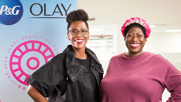 Two women with black hair are standing together and smiling in front of a poster that has the Olay and P&G logos on it.