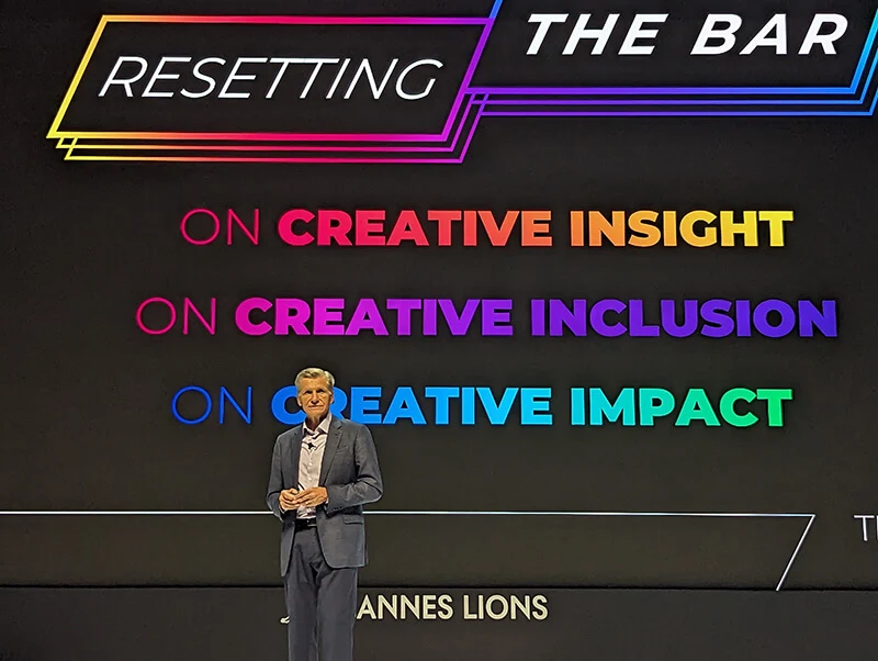 A tall, white male with short gray hear, wearing a grey suit and collared shirt, stands on a stage with a backdrop of colorful taglines about Procter & Gamble's approach to creativity.