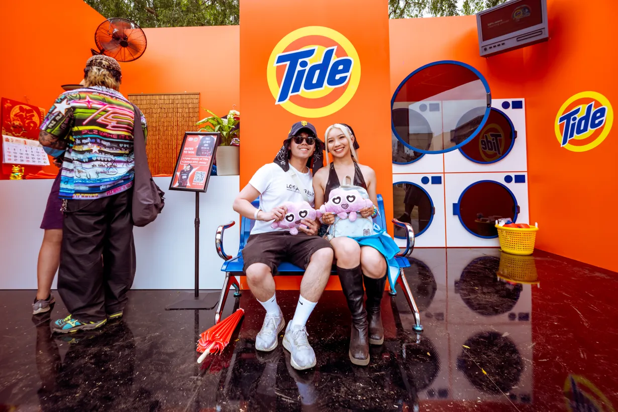 Two people seated on a bench holding plush toys at a Tide-branded event with washing machines and bright orange displays in the background.