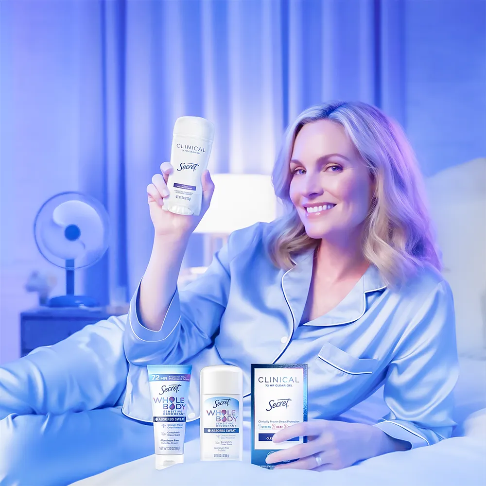 A blonde middle-aged woman lies on a bed wearing lavender silk pajamas. She holds up a stick of deodorant. The room is softly lit in a matching lavender tone.