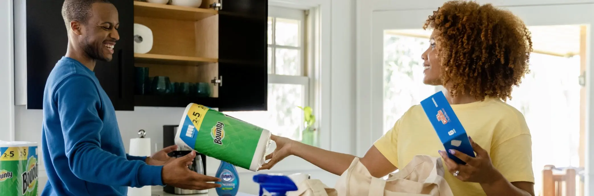 Woman and man are unpacking groceries in kitchen. Woman hands man a roll of Bounty paper towels.