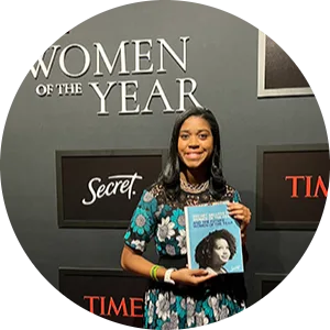 Smiling Black woman standing in front of a 'Women of the Year' PR wall and looking at the camera