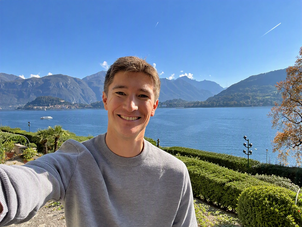 A smiling man in a grey sweater stands outdoors on a sunny day. A calm lake and distant mountains sit behind him.