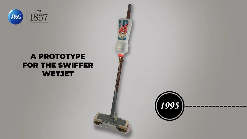 A simple Swiffer Wetjet floor cleaning apparatus is photographed against a white backdrop. Black text reads, "A prototype for the swiffer wet jet. 1995."