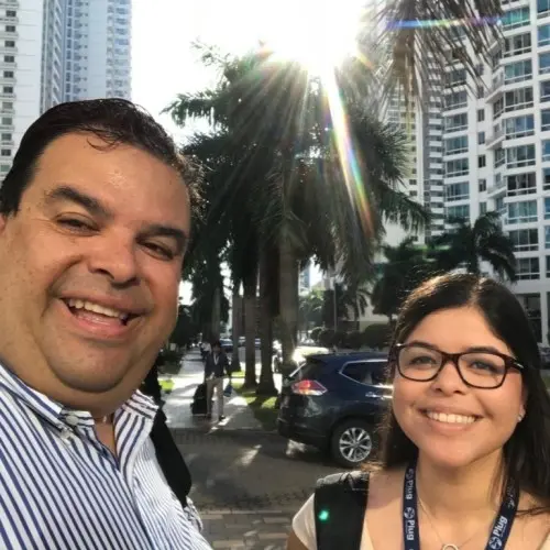 A Hispanic male with short dark hair and a young Hispanic women with long dark hair and black-framed glasses smile as they stand on city street. Skyscrapers and palm trees are in the background.