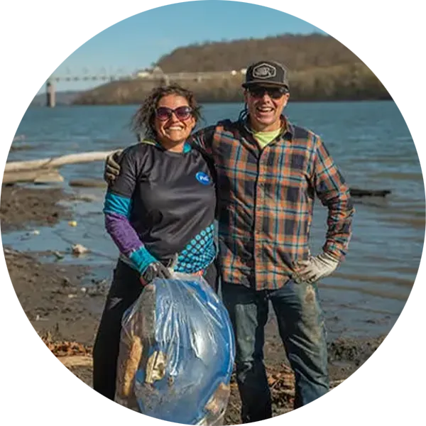 Two volunteers standing on a shoreline during an Ohio River Clean Up, holding a large bag of collected waste with water and a bridge in the background.