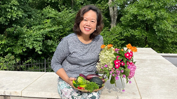 An Asian woman with shoulder length dark hair wears a black and white top. She smiles as she sits outside, while holding a basket of vegetables and a bouquet of flowers.
