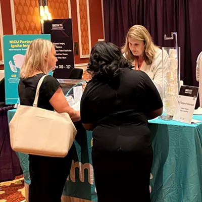 Three women interact at a conference booth with a teal tablecloth. Two women face away from the camera, while a third assists them behind the table.
