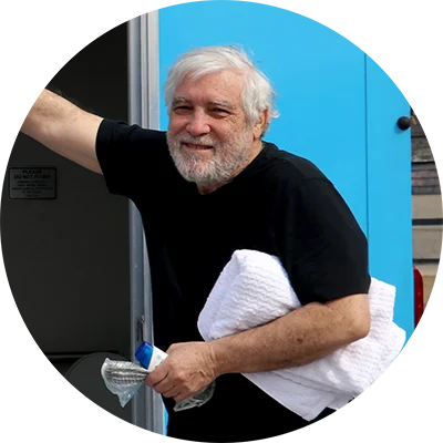 Person with gray hair wearing a black T‑shirt stands at the open door of a blue and gray vehicle, holding a white folded towel and a plastic water bottle.