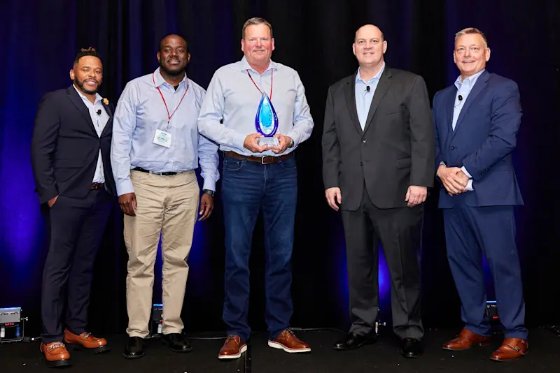 Five men stand side by side on a stage with a dark backdrop. The man in the center holds up a glass trophy.