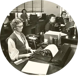 A black and white photo of P&G’s first blind employee, Jane Dotson, who was hired in January 1950 and worked in the transcribing department at the General Offices. She sits at a desk, typing on a typewriter amongst other women transcribers.