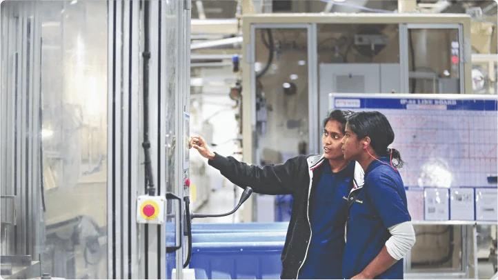 Two women are standing in a plant and are facing a glass room.
