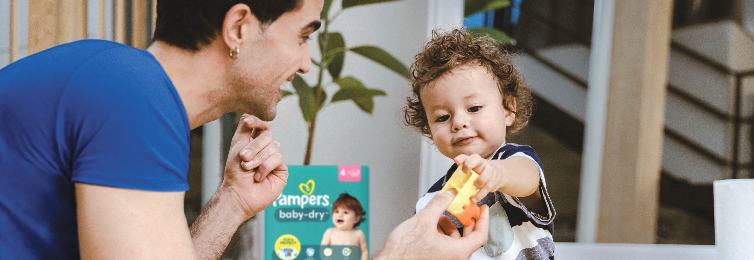 Man on floor playing with baby, handing the baby a toy car. A pack of Pampers diapers is in the background.  