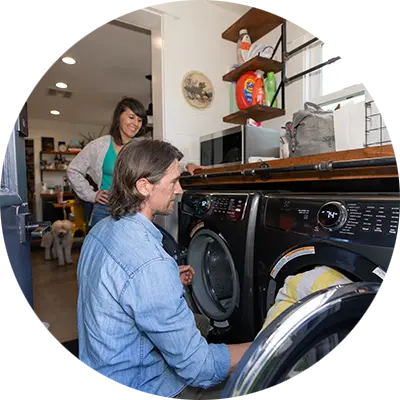 A man with long hair and a blue shirt removed laundry from the dryer as a smiling woman looks on. A container of Tide Pods can be seen on a shelf above them.