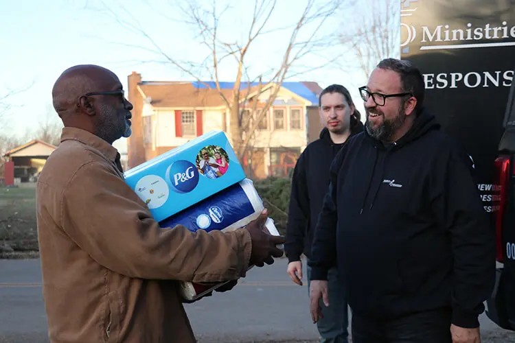 A man holds a cardboard P&G box and Charmin toilet paper as he smiles at a man. In the background are another man, a black truck and a two story house.