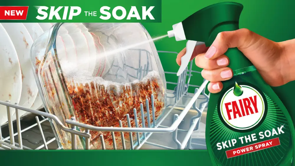 A close up of someone spraying a glass baking dish that has baked-on food residue with a bottle of "Fairy Skip The Soak Power Spray". The words “New” and “Skip the Soak” are prominently displayed at the top of the image.