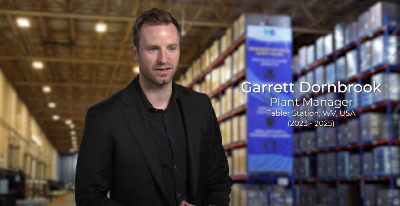 A man in a black suit standing in front of a large manufacturing plant.