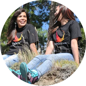 Two women in black t-shirts are sitting in grass and smiling towards each other.