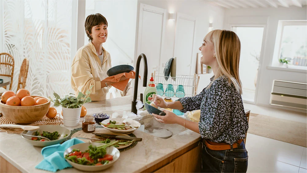 Two women smile while doing dishes – one is washing, the other drying.  