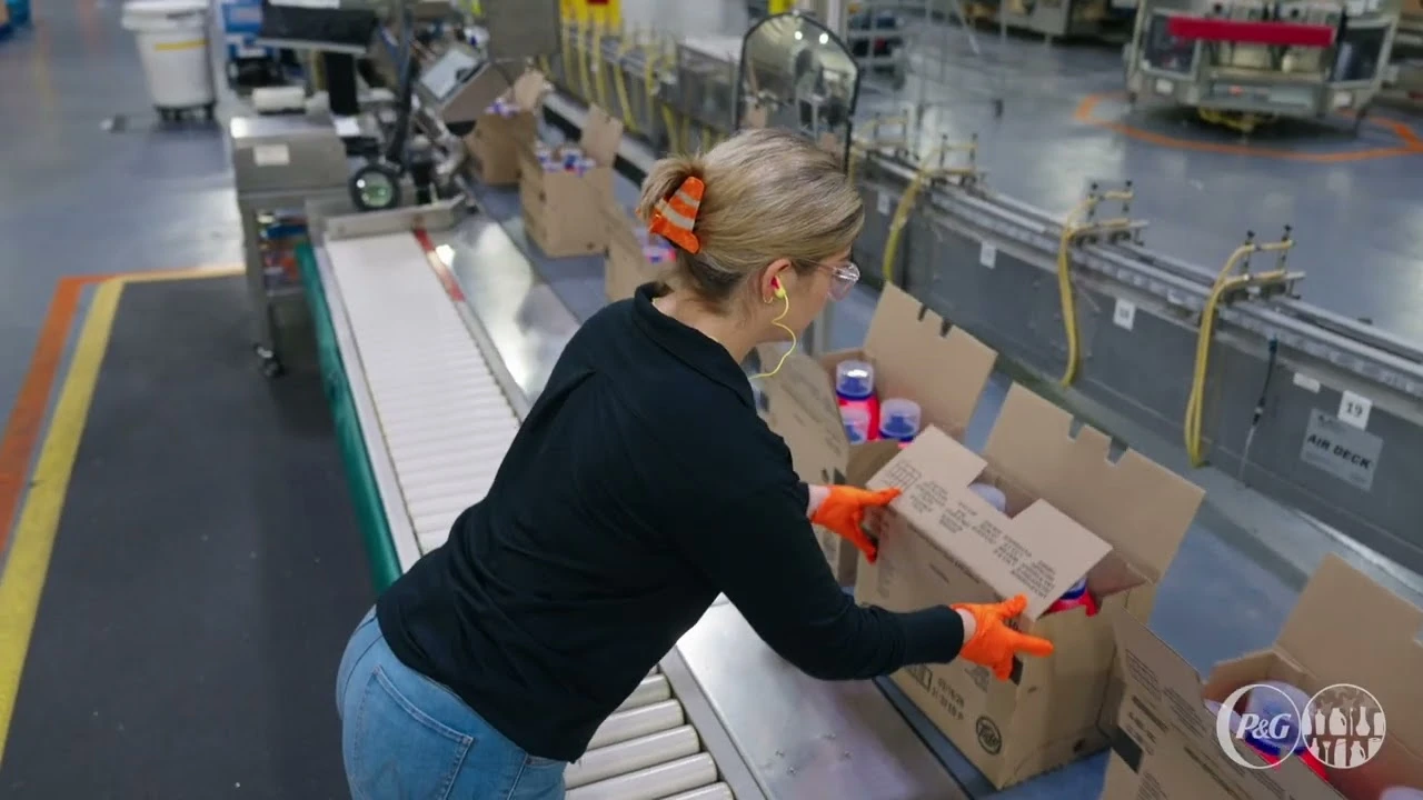 Worker wearing gloves and safety glasses packing products into cardboard boxes on a conveyor line in a manufacturing facility.