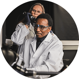 A man and woman in white lab coats. The woman holds up a tool to the man's head for a hair analysis.