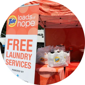 Multiple white tote bags with the Tide logo are on top of a table with an orange tablecloth. They are next to a sign that says “Tide loads of hope: Free Laundry Services.” 