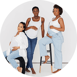 Three Black women in white shirts and blue jeans. All three women are pregnant and pose while standing or sitting on stools.
